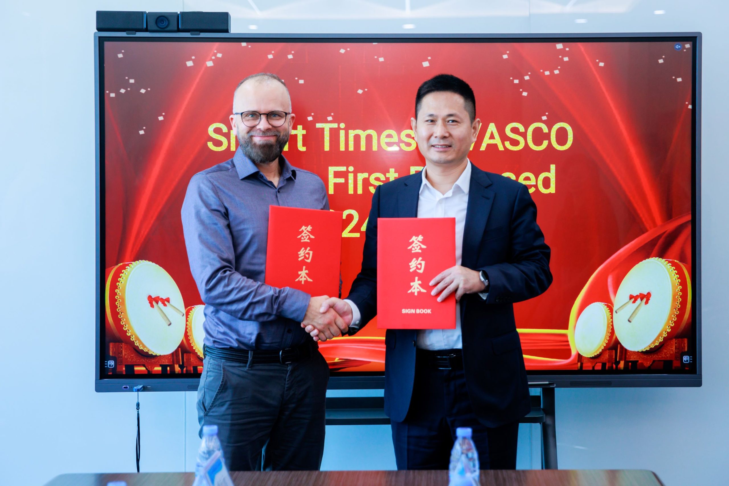 Zwei Männer geben sich vor einem Bildschirm mit der Aufschrift „Smart Times / VASCO“ die Hand und halten rote Mappen mit chinesischer Beschriftung. Das Foto wurde bei einer offiziellen Veranstaltung anlässlich des Markteintritts von Vasco in den chinesischen Markt aufgenommen.