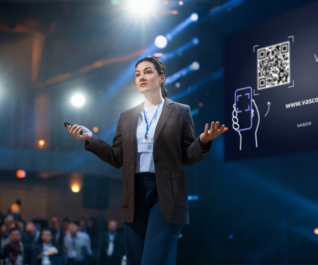 Eine Frau hält einen Vortrag auf einer Konferenz. Hinter ihr auf dem Bildschirm ist der Anmeldebildschirm für Vasco Audience mit einem QR-Code zu sehen.