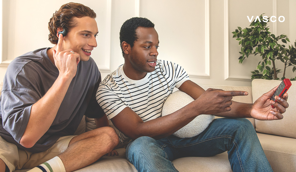 Zwei junge Männer unterhalten sich. Einer trägt einen Vasco E1 Übersetzungskopfhörer, der andere hat einen tragbaren Vasco Translator Q1 in der Hand und zeigt auf den Bildschirm.
