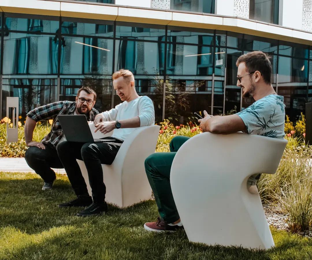 Three men sit on modern white chairs outside a building, working together on a laptop. They appear relaxed and engaged, with one man smiling while looking at his translator. Lush grass and flowers surround them.