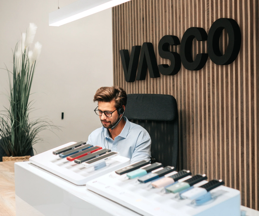 Ein Mann vom Kundendienst sitzt an einem Schreibtisch, an dem mehrere Vasco Translator-Geräte in verschiedenen Farben ausgestellt sind, hinter ihm hängt ein großes