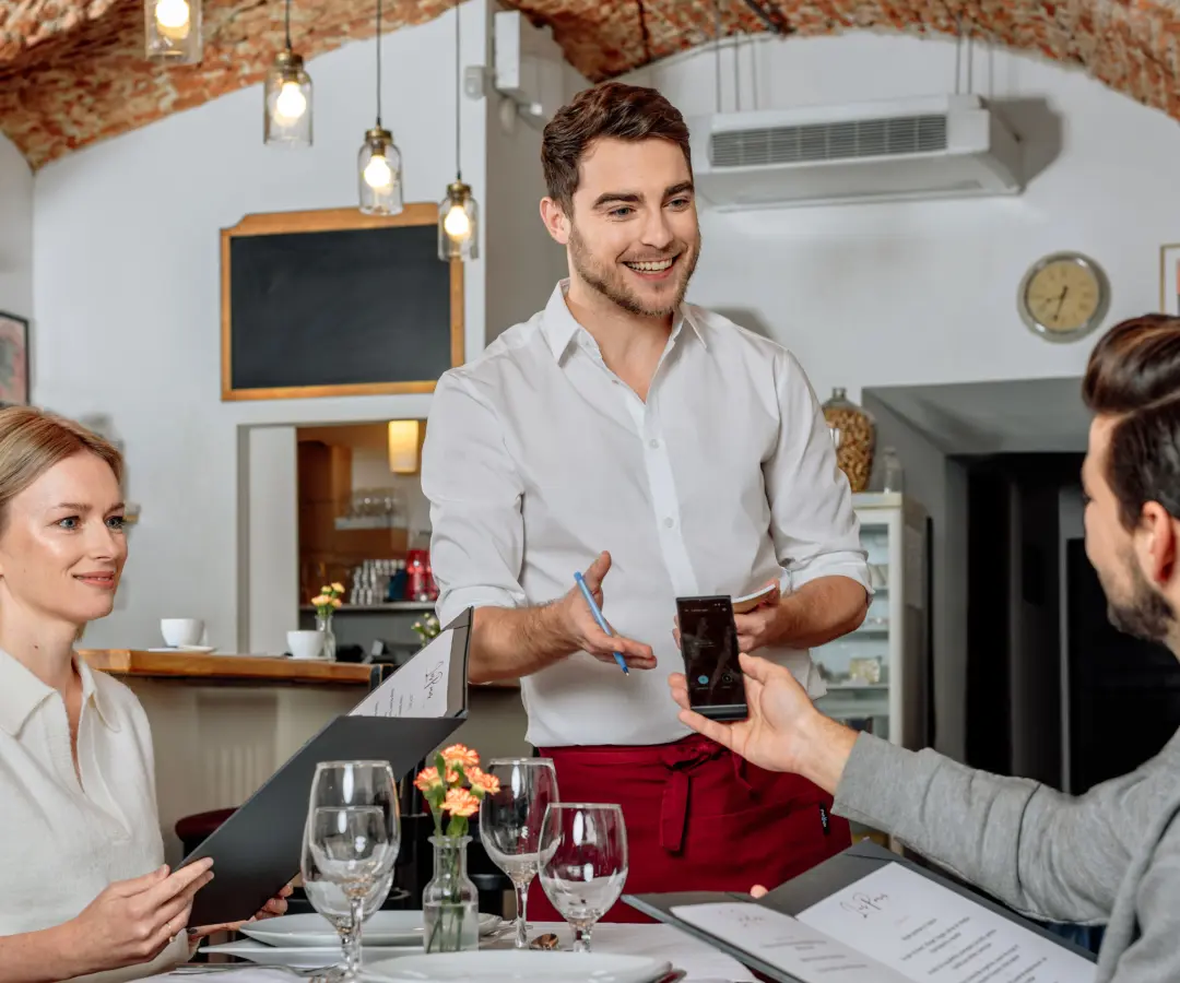 Ein lächelnder Kellner steht an einem Restauranttisch, während ein sitzender männlicher Kunde einen Übersetzer in der Hand hält. Eine Kundin sitzt in der Nähe und hält eine Speisekarte in der Hand. Der Tisch ist mit Gläsern, Tellern und einer kleinen Blumenvase gedeckt.
