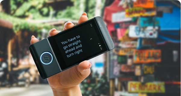 Nahaufnahme einer Hand, die einen schwarzen elektronischen Übersetzer der Marke Vasco waagerecht hält. Der Bildschirm ist im Vollbildmodus und zeigt den übersetzten Text in einer großen, klaren blauen Schrift an: „Sie müssen geradeaus fahren und dann rechts abbiegen.“ Im Hintergrund ist eine lebhafte, unscharfe Außenaufnahme zu sehen, auf der ein bunter Pfosten zu sehen ist, der mit Wegweisern für verschiedene Länder wie Polen, Chile und Dänemark bedeckt ist. Das Sonnenlicht ist hell und hebt den Text auf dem Gerät hervor, sodass er gut lesbar ist. 