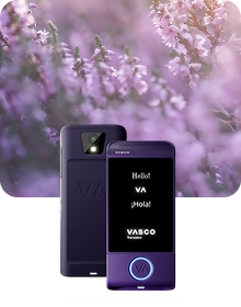 Ein Produktpräsentationsbild, das ein violettes „Vasco Translator“-Gerät aus zwei Blickwinkeln vor einem dunklen, strukturierten Stoffhintergrund zeigt. Das Gerät auf der linken Seite ist von hinten zu sehen, wobei eine Kameralinse mit Blitz und ein geprägtes „VA“-Logo hervorgehoben sind. Das Gerät auf der rechten Seite zeigt die Vorderansicht mit beleuchtetem Bildschirm, auf dem die Begrüßungen „Hello!“, „VA“, „¡Hola!“ und das „VASCO Translator“-Logo zu sehen sind. Ein runder Knopf am unteren Rand der Vorderseite des Geräts ist von einem leuchtend blauen Licht umrandet. Die Komposition ist vor einem verträumten, unscharfen Hintergrund aus violetten Heidekrautblüten vor dem Hintergrund von sanftem Sonnenlicht angeordnet.