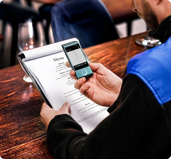 Eine Nahaufnahme aus der Vogelperspektive einer Person, die an einem Holztisch in einem Restaurant sitzt und ein elektronisches Übersetzungsgerät benutzt, um eine Speisekarte zu lesen. Die Person hält eine weiße Papierspeisekarte in der linken Hand, während sie mit der rechten Hand ein kleines, blaugrünes Vasco-Übersetzungsgerät darüber hält. Auf dem Bildschirm des Geräts ist eine digitale Version der Speisekarte zu sehen, die ins Polnische übersetzt wird. Die Person trägt eine schwarze Jacke mit einem hellblauen Schulterbesatz. Der Hintergrund ist sanft unscharf und zeigt das gemütliche Interieur eines Restaurants. 