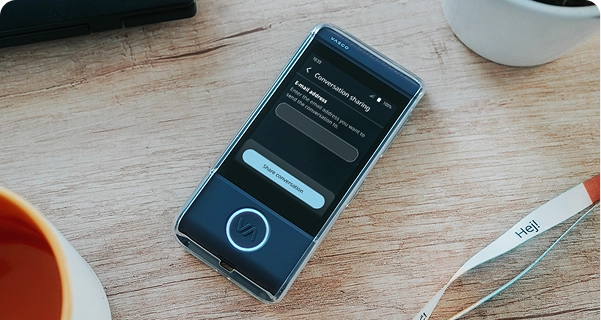 Eine Nahaufnahme aus der Vogelperspektive eines dunkelblauen elektronischen Übersetzers der Marke Vasco in einer durchsichtigen Schutzhülle, der auf einem hellen Holztisch liegt. Der Bildschirm des Geräts ist aktiv und zeigt ein Menü „Konversation teilen“ mit einem Feld zur Eingabe einer E-Mail-Adresse und einer Schaltfläche mit der Aufschrift „Konversation teilen“. Um das Gerät herum liegen auf dem Tisch der Rand einer orangefarbenen Kaffeetasse, ein weißer Blumentopf und ein weißes Schlüsselband mit der Aufschrift „Hej!“. Die Szene ist hell beleuchtet, was eine klare und moderne Ästhetik erzeugt. 