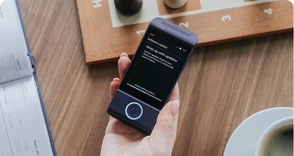 Eine Aufnahme von oben auf die Hand einer Person, die einen violetten elektronischen Übersetzer der Marke Vasco über einem Holztisch hält. Auf dem Bildschirm des Geräts ist die Meldung „Software-Update“ zu sehen, die lautet: „Bleiben Sie auf dem Laufenden“, sowie ein Fortschrittsbalken mit dem Text „Update wird heruntergeladen...“. Auf dem Tisch um das Gerät herum liegen ein hölzernes Schachbrett mit Schachfiguren, ein aufgeschlagener Kalender und eine weiße Tasse Kaffee. Die Beleuchtung ist sanft und natürlich und schafft eine entspannte, alltägliche Atmosphäre. 