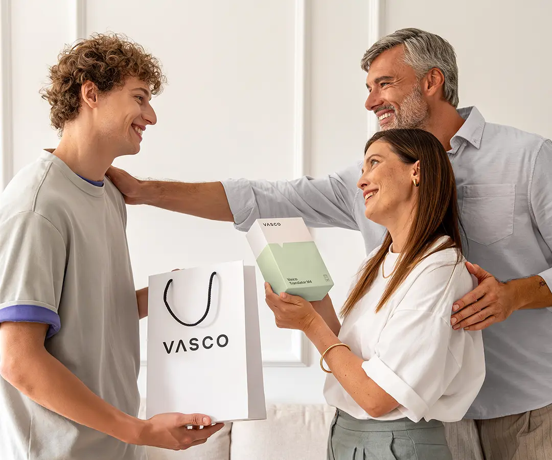 Eine herzerwärmende Szene, in der drei Menschen lächelnd beieinander stehen. Links hält ein junger Mann mit lockigem Haar eine weiße „VASCO“-Einkaufstüte in der Hand. In der Mitte hält eine Frau eine weiß-hellgrüne Schachtel mit der Aufschrift „Vasco Translator M4“ und blickt den jungen Mann glücklich an. Hinter ihr steht ein Mann mit graumeliertem Haar, der warmherzig lächelt, wobei er eine Hand auf die Schulter der Frau und die andere auf die Schulter des jungen Mannes legt. Sie befinden sich in einem hellen Raum mit einer Wand in neutralen Farbtönen und einem Sofa im Hintergrund. 