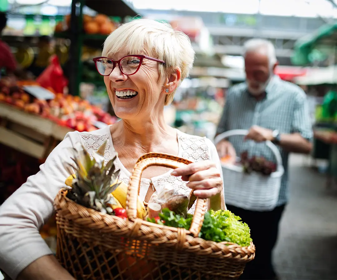 Eine lächelnde ältere Frau mit kurzen blonden Haaren und einer roten Brille, die auf einem Markt im Freien einen mit frischem Obst und Gemüse gefüllten Weidenkorb trägt. Im unscharfen Hintergrund ist eine weitere Person zu sehen, die zwischen den Ständen mit Obst und Gemüse einkauft.