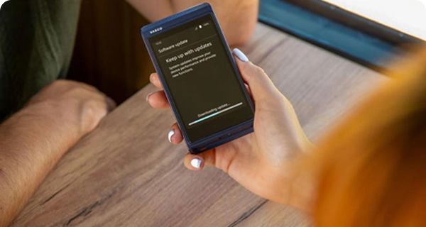 Eine Frau hält in einem Zug über einen Holztisch hinweg einen blauen Vasco Translator Q1. Auf dem Bildschirm des Geräts wird eine Benachrichtigung über ein Software-Update mit einem Fortschrittsbalken angezeigt, der „Update wird heruntergeladen…“ anzeigt.